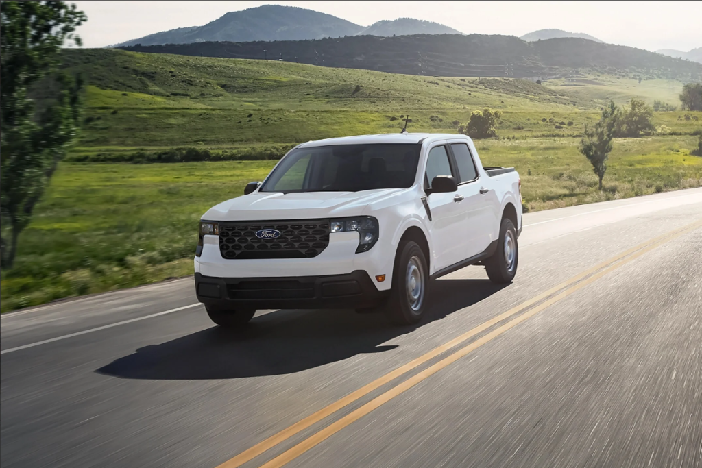 A white 2025 Ford Maverick driving down a country road with grass fields in the backround.